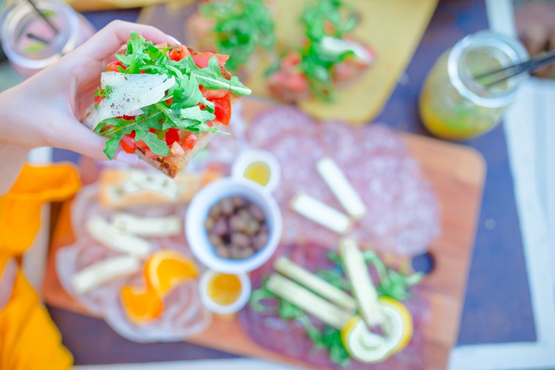 La foto muestra en primer plano una mano femenina sosteniendo una bruscheta con tomates, rúcula y queso. En el fondo, desenfocado, se ven bebidas y otros platos ideales para una cena de verano en terraza