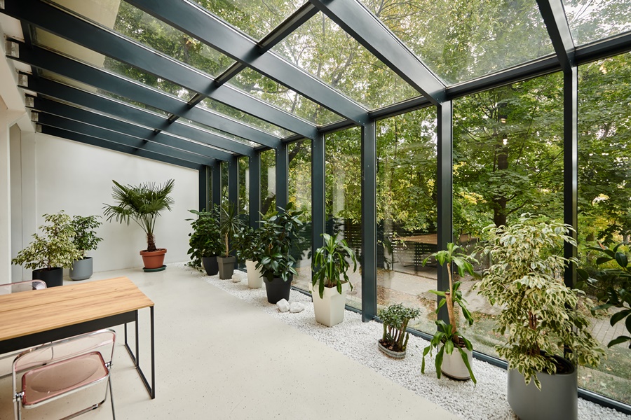 Galería acristalada orientada al jardín con estructura metálica y plantas decorativas, diseñada para ahorrar energía con cerramiento de cristal mediante aprovechamiento solar.