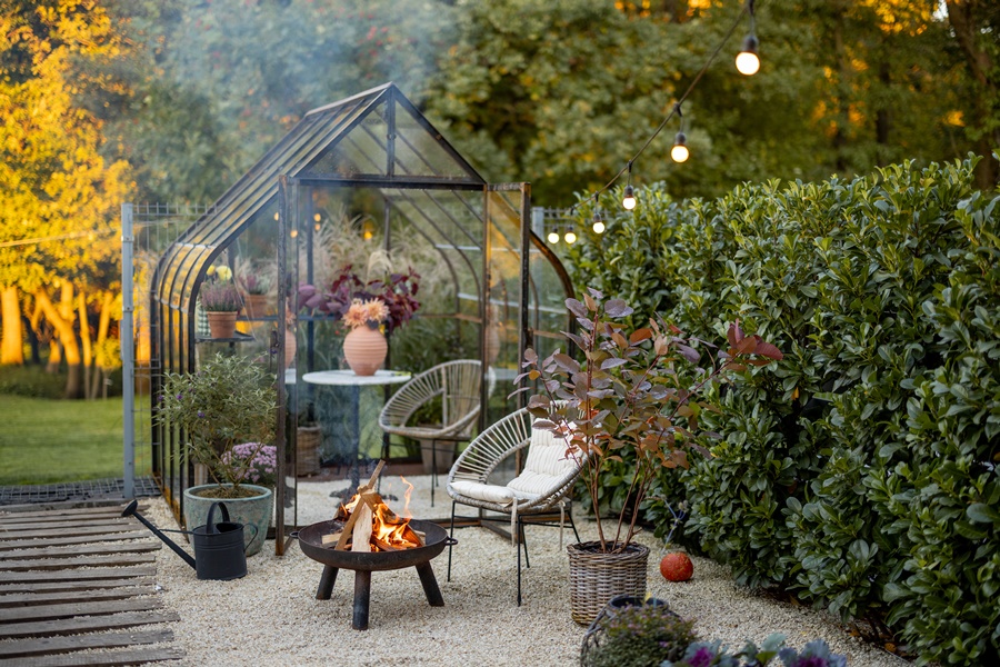 Cerramiento de cristal tipo invernadero en jardines pequeños y sencillos, rodeado de vegetación, sillas de mimbre y zona de fuego para crear un rincón de relax funcional y estético.