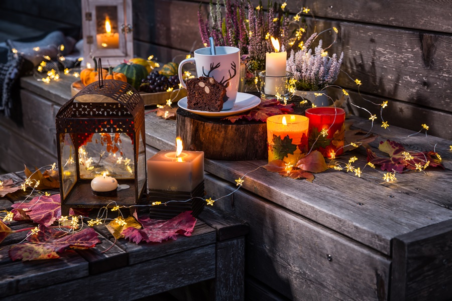 Decoración exterior terraza con velas encendidas, hojas secas, guirnaldas luminosas y detalles otoñales sobre banco de madera con bebida caliente y bizcocho.